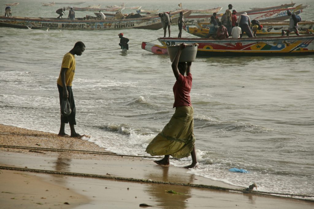 Banjul, The Gambia, April 1, 2008, a woman is carrying a bucket full of fish on her head from the fisher boats, where many people are working, beside a man is looking the water