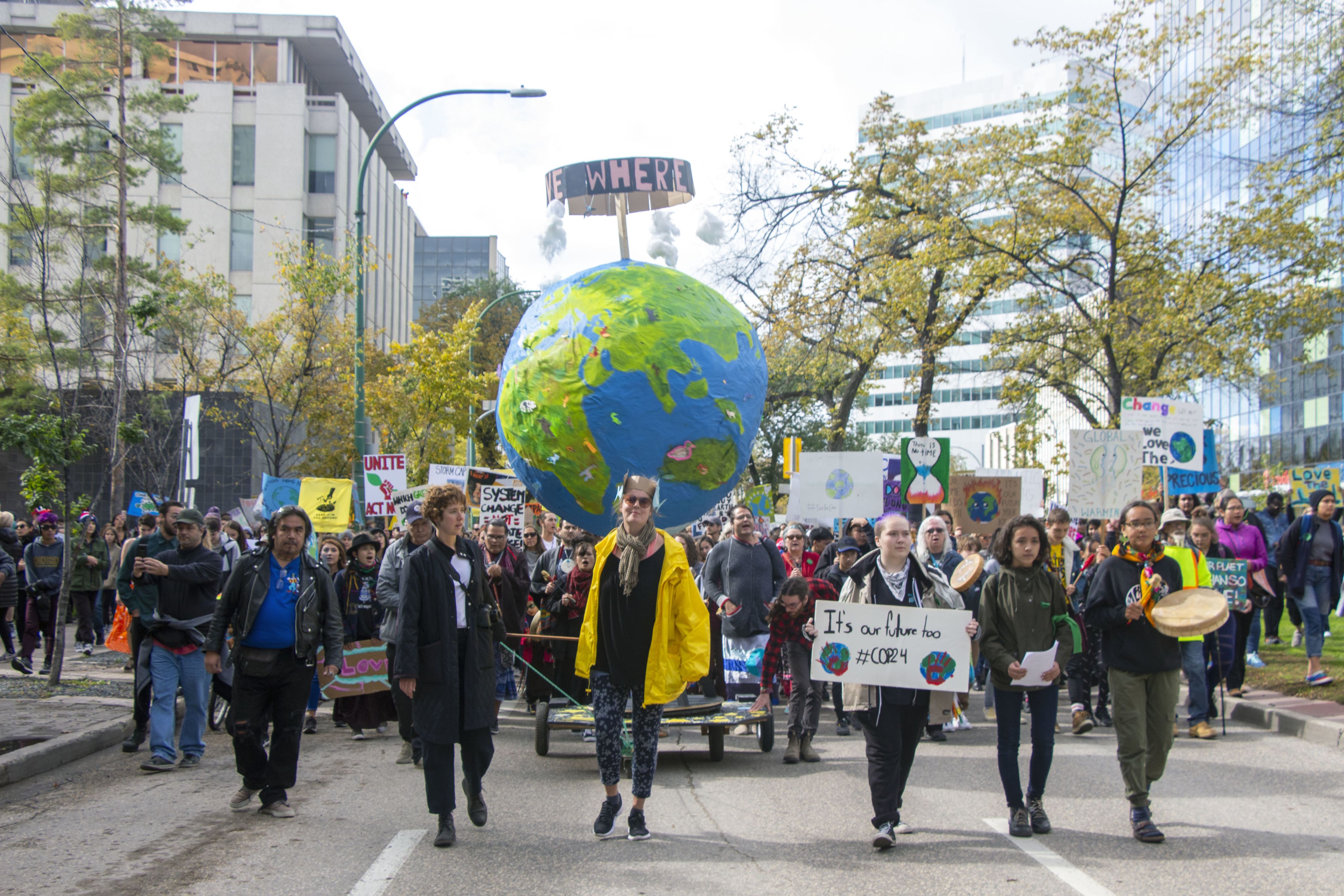 Front line of a climate march walks down the street in Winnipeg in front of a large globe and thousands of people following them.