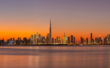 Panorama of Dubai Business Bay skyline at night after sunset with colorful illuminated buildings and calm Dubai Creek water.