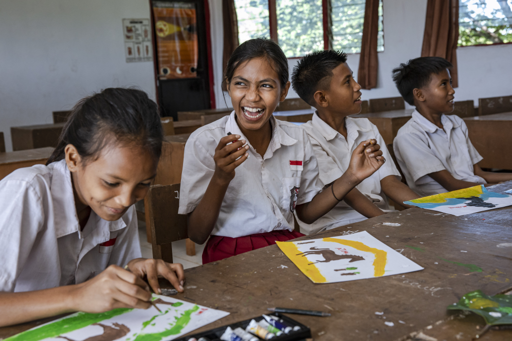 Three students in white and red uniforms sit at a long wooden desk in a bright classroom, engaged in an art activity. In the center, a girl laughs joyfully while holding a small paint tube.