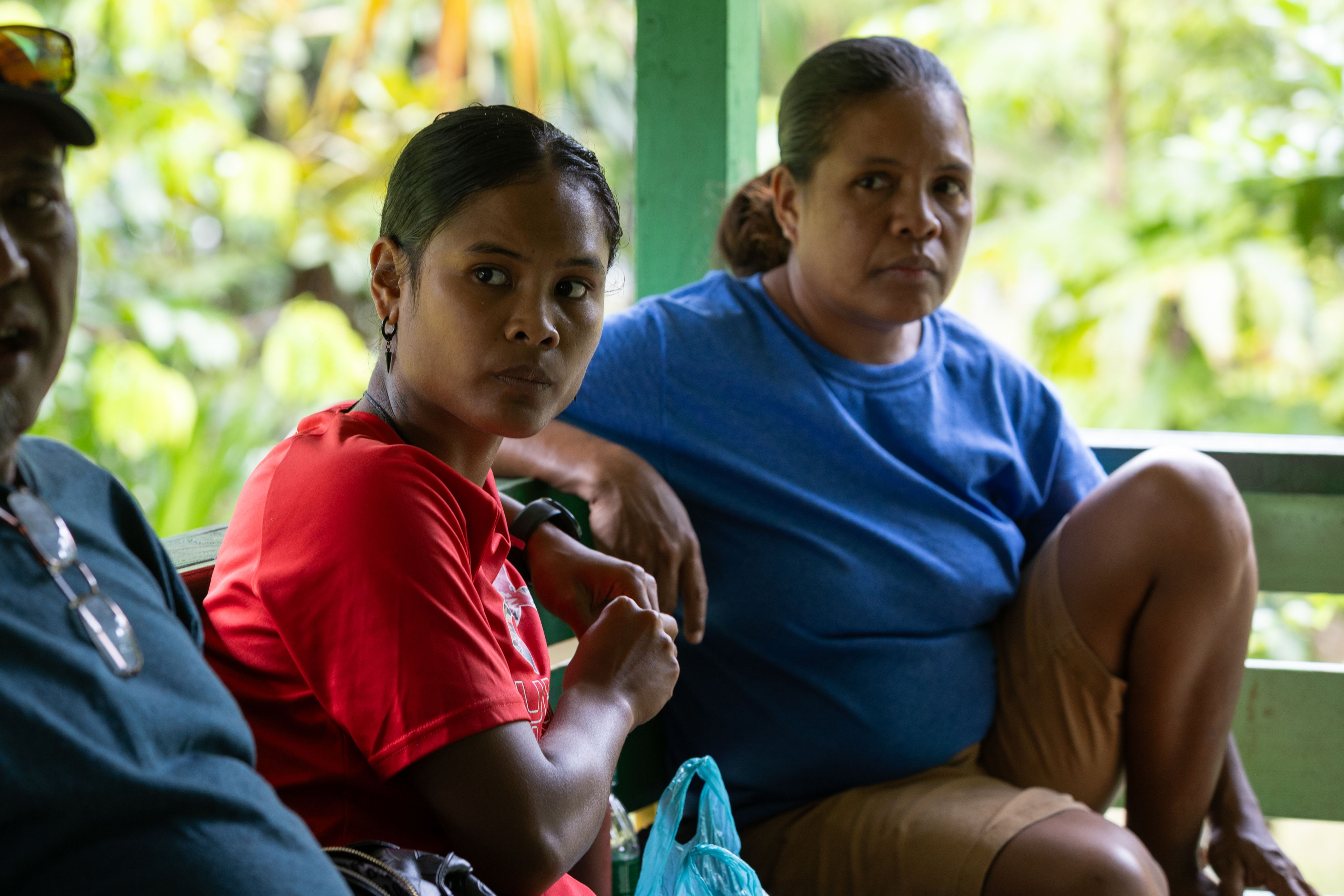 A woman in a blue shirt and a woman in a red shirt sit on a bench outside. 
