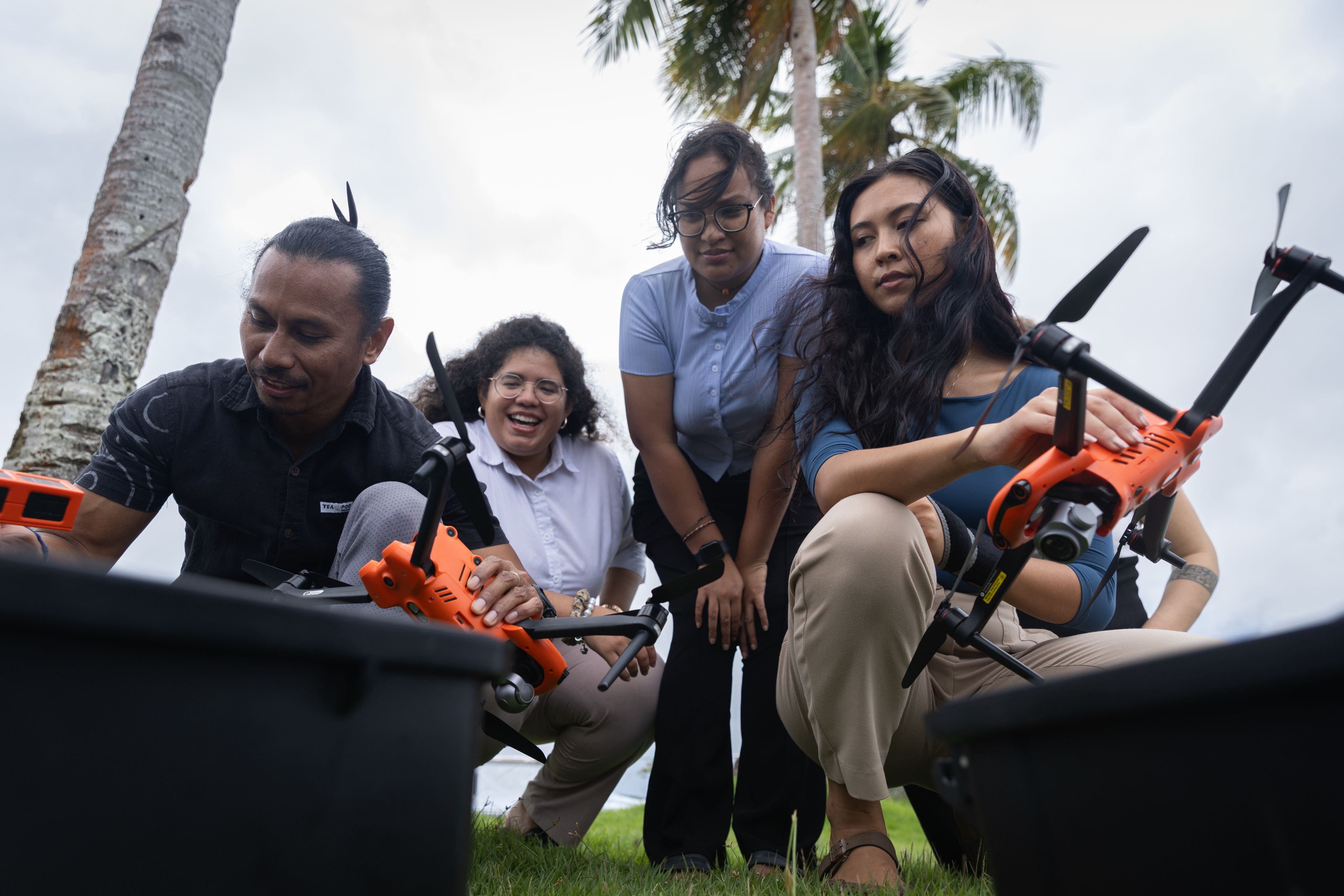 Four people crouching outside on a cloudy day. Two in the front hold drones. 