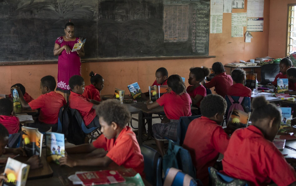 A teacher stands at the front of a classroom reading from a booklet while young students in red uniforms sit at their desks following along with their own books.